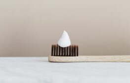 En trätandborste med mjuka, bruna borst har en klick vit tandkräm på, placerad på en vit marmoryta med en enkel beige bakgrund, redo för ditt nästa besök hos tandläkare Eskilstuna.