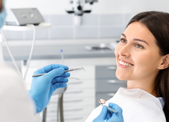 En kvinna sitter i en tandläkarstol och ler mot en tandläkare Eskilstuna som håller i tandinstrument. Hon har en vit haklapp på sig och tandläkaren har blå handskar och mask. Bakgrunden visar tandläkarutrustning.